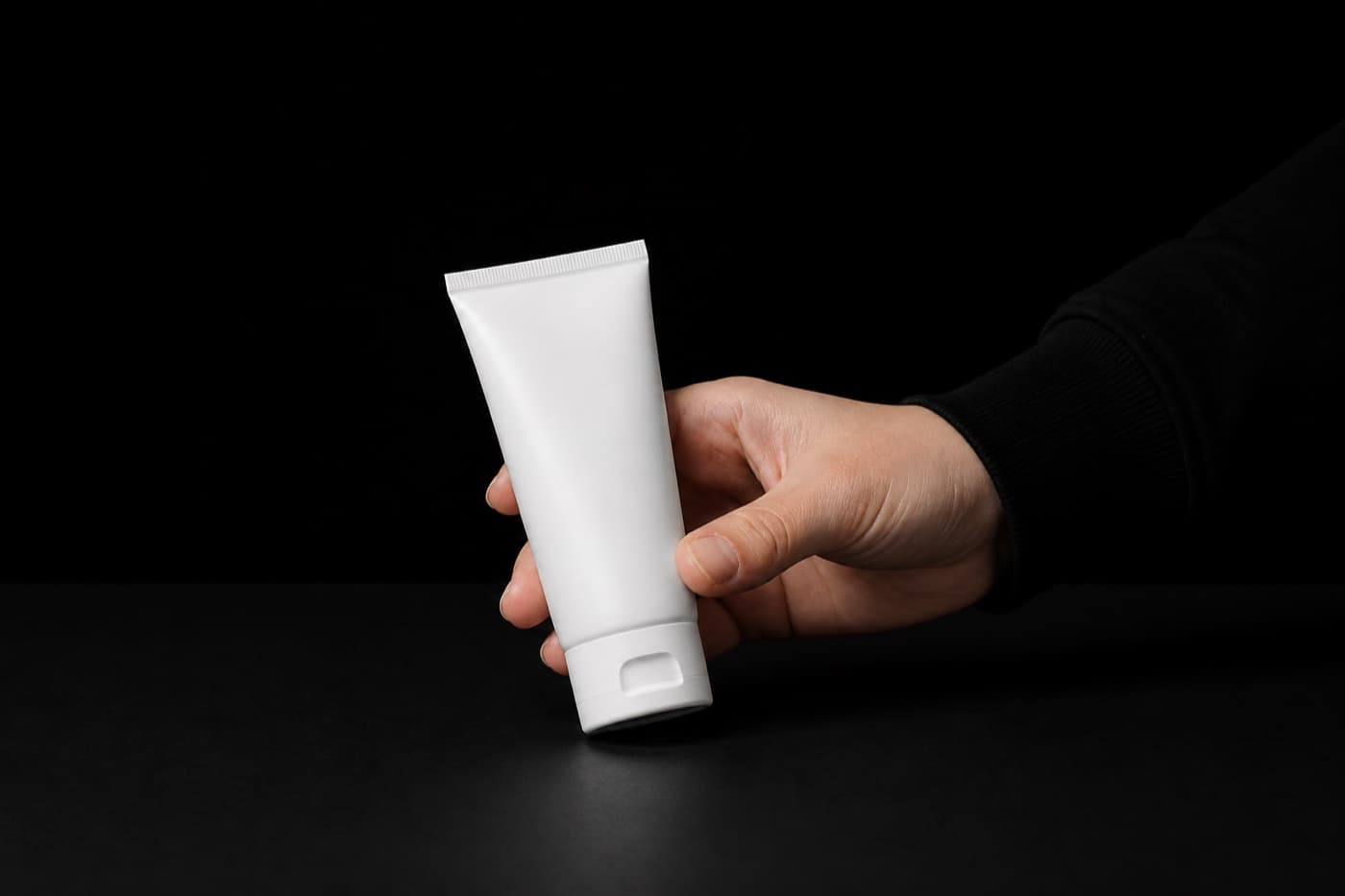 검은 테이블 위에서 검정 긴소매를 입은 손 하나만 프레임 오른쪽에서 들어와 무지 화장품 튜브를 집어 들고 있다